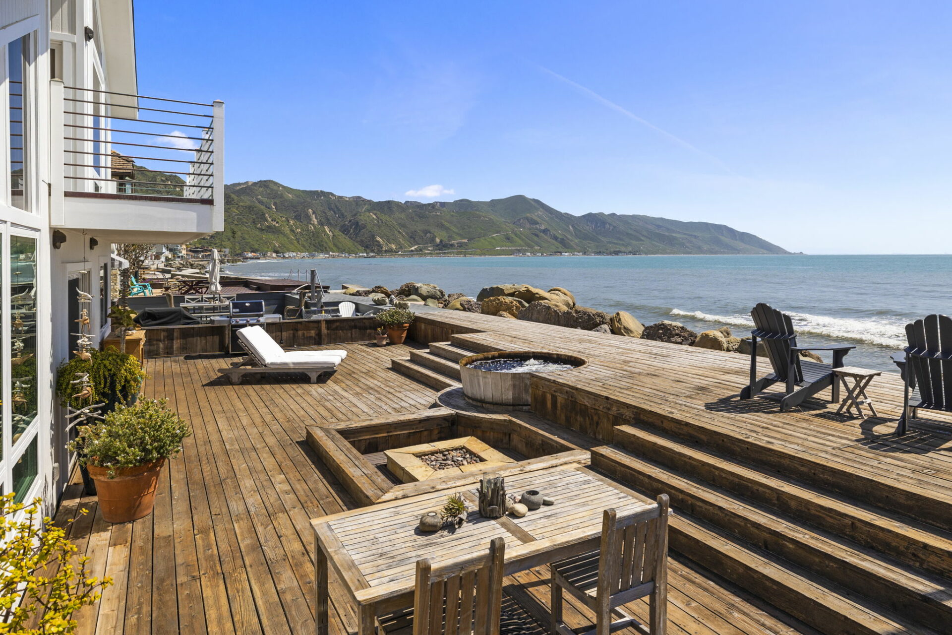 Coastal terrace with outdoor furniture overlooking a scenic view of the ocean and hills.